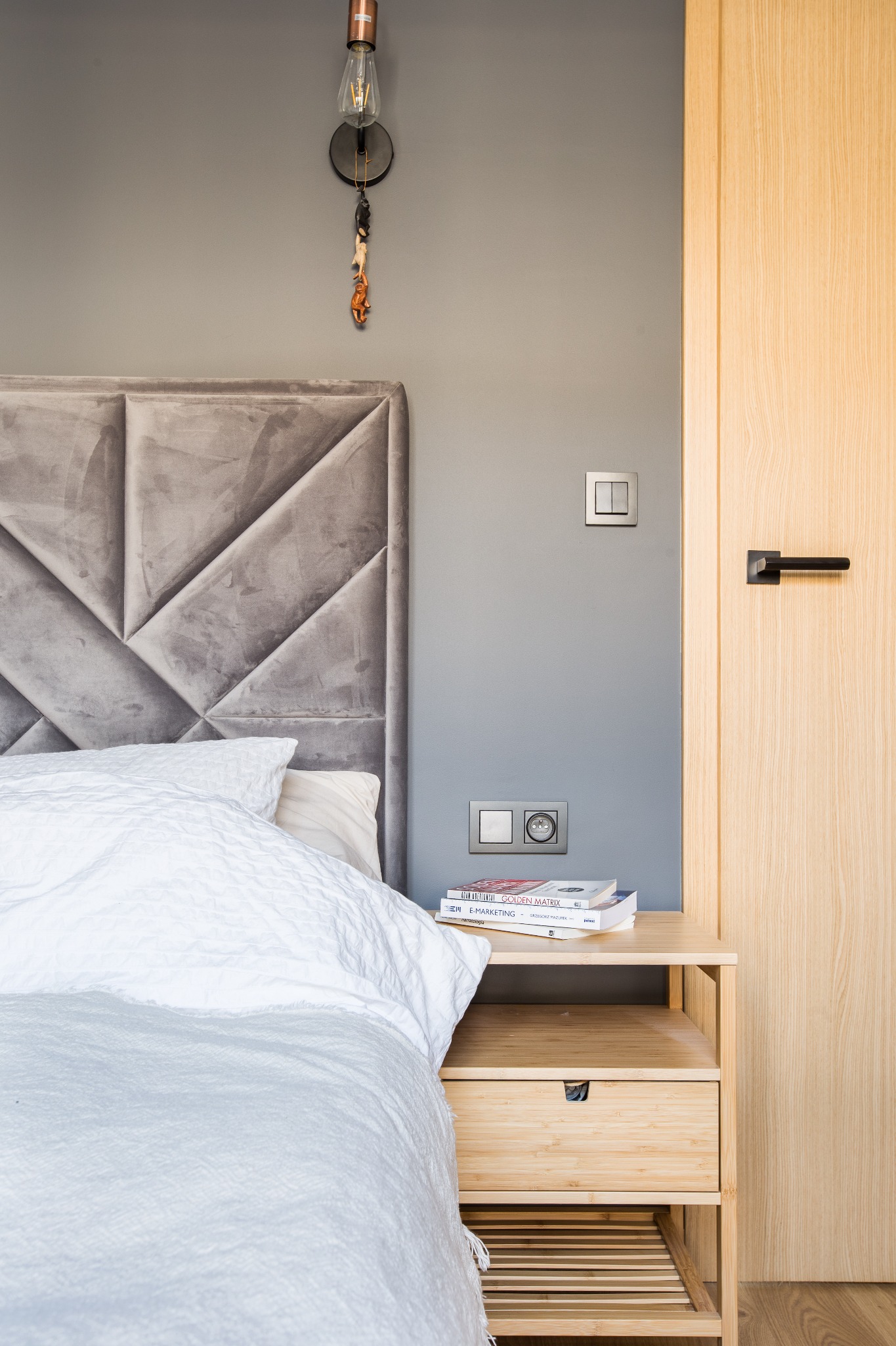 Sypialnia z szarą tapicerowaną ścianą za łóżkiem, drewniana szafka nocna z książkami i lampa wisząca z ozdobną figurką.