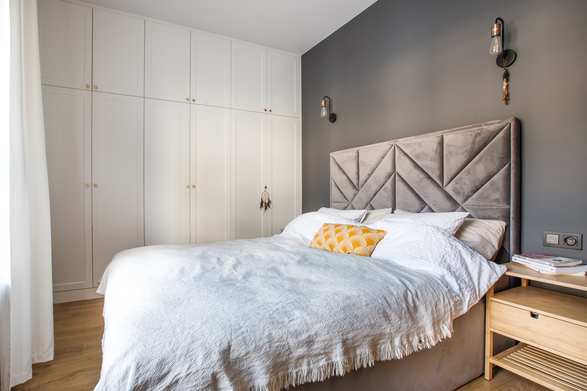 Nowoczesna sypialnia z szarą ścianą, tapicerowanym zagłówkiem w geometryczne wzory, białą pościelą z żółtą poduszką i szafką nocną z książkami.
