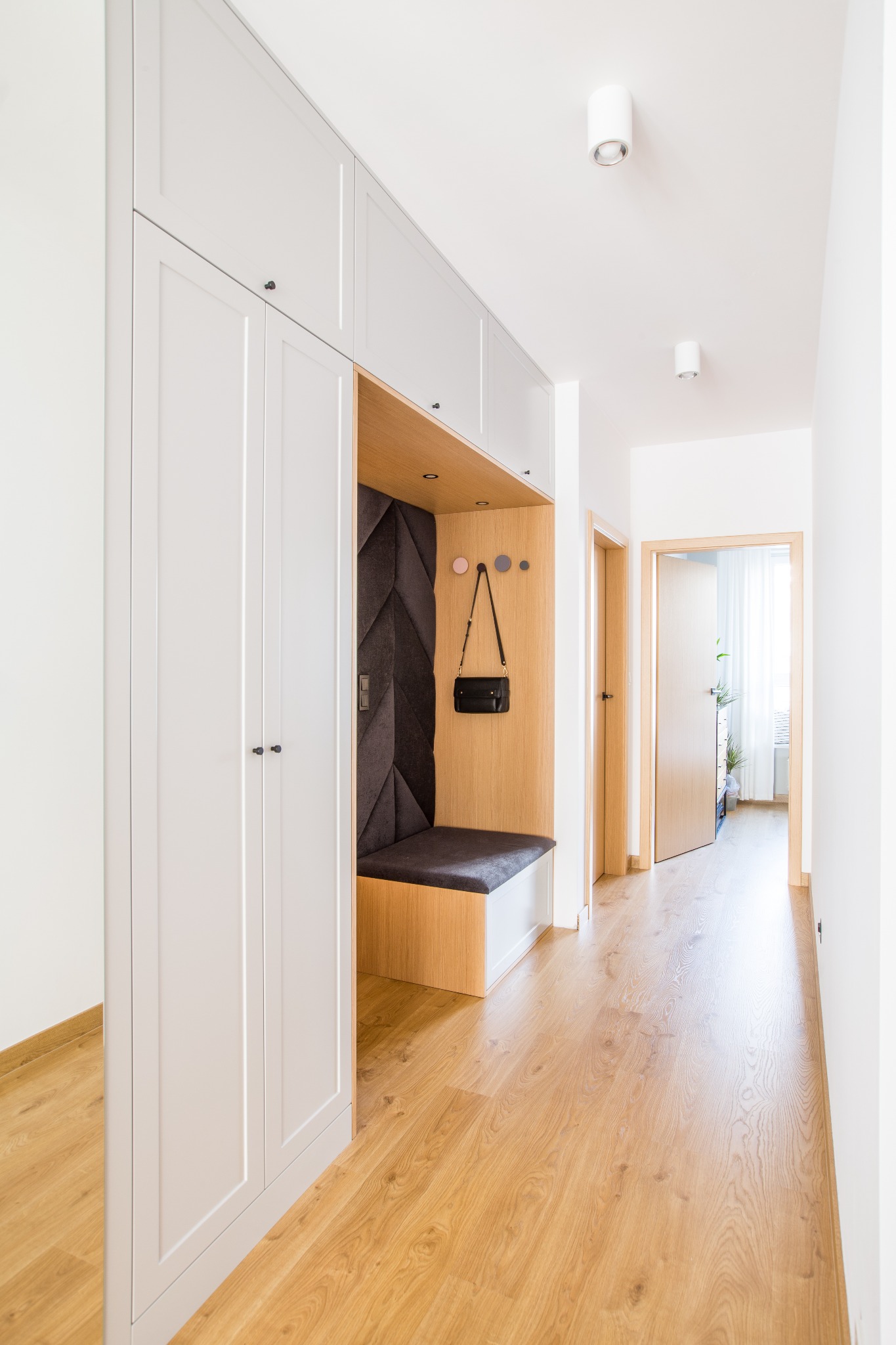 Nowoczesny przedpokój z zabudową meblową w kolorze jasnego drewna i szarości, wnęka z siedziskiem i tapicerowanym panelem ściennym w geometryczny wzór, wisząca torebka, drewniana podłoga i białe...