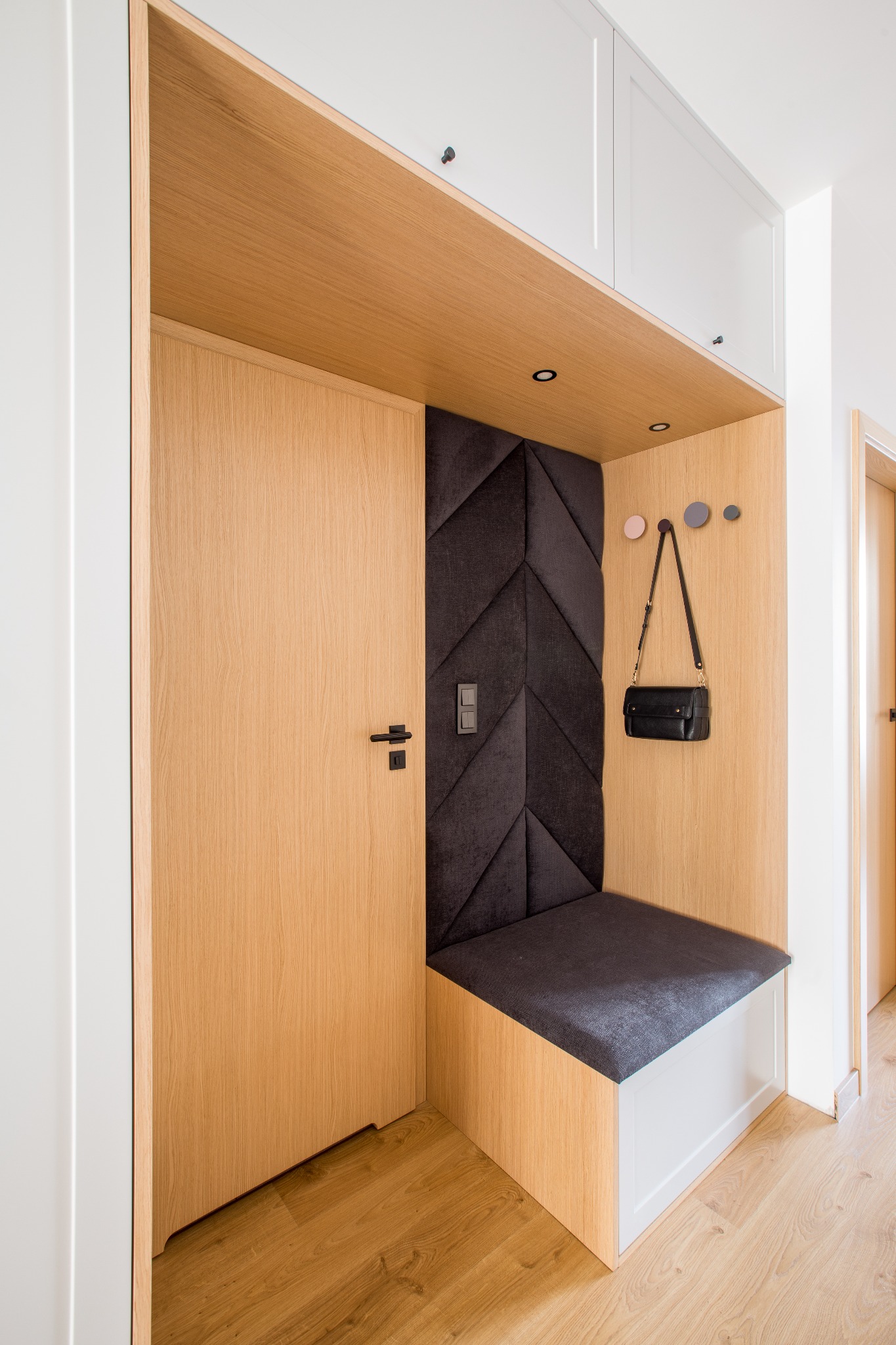 Nowoczesna garderoba w przedpokoju z drewnianymi elementami, tapicerowanym siedziskiem i panelem ściennym z geometrycznym wzorem, oświetleniem punktowym i czarną torebką wiszącą na haczyku.