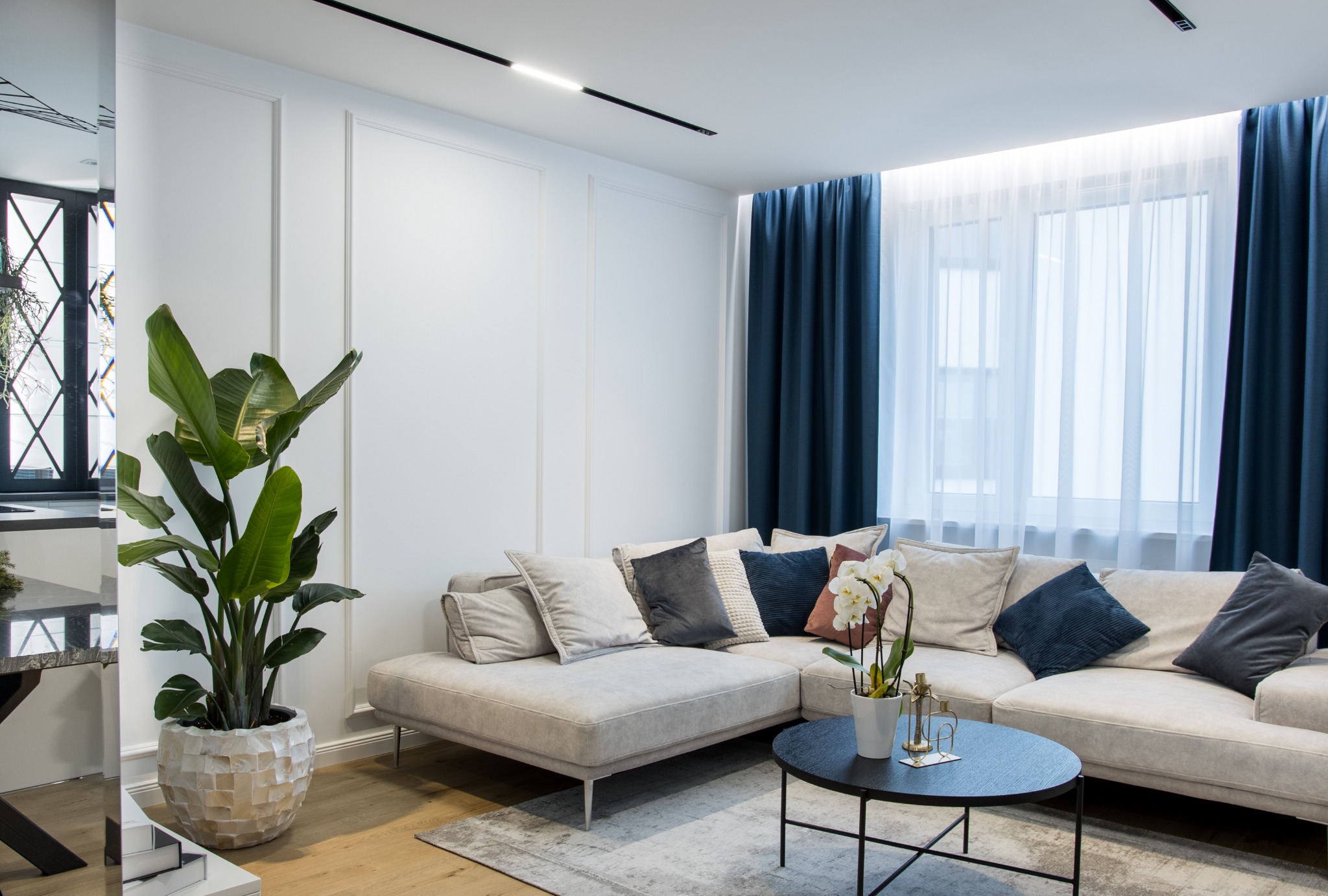 Jasny salon z narożną sofą w odcieniach beżu, dekoracyjnymi poduszkami, okrągłym czarnym stolikiem kawowym i dużą rośliną doniczkową w ozdobnej osłonce, doświetlony oknem z granatowymi zasłonami.