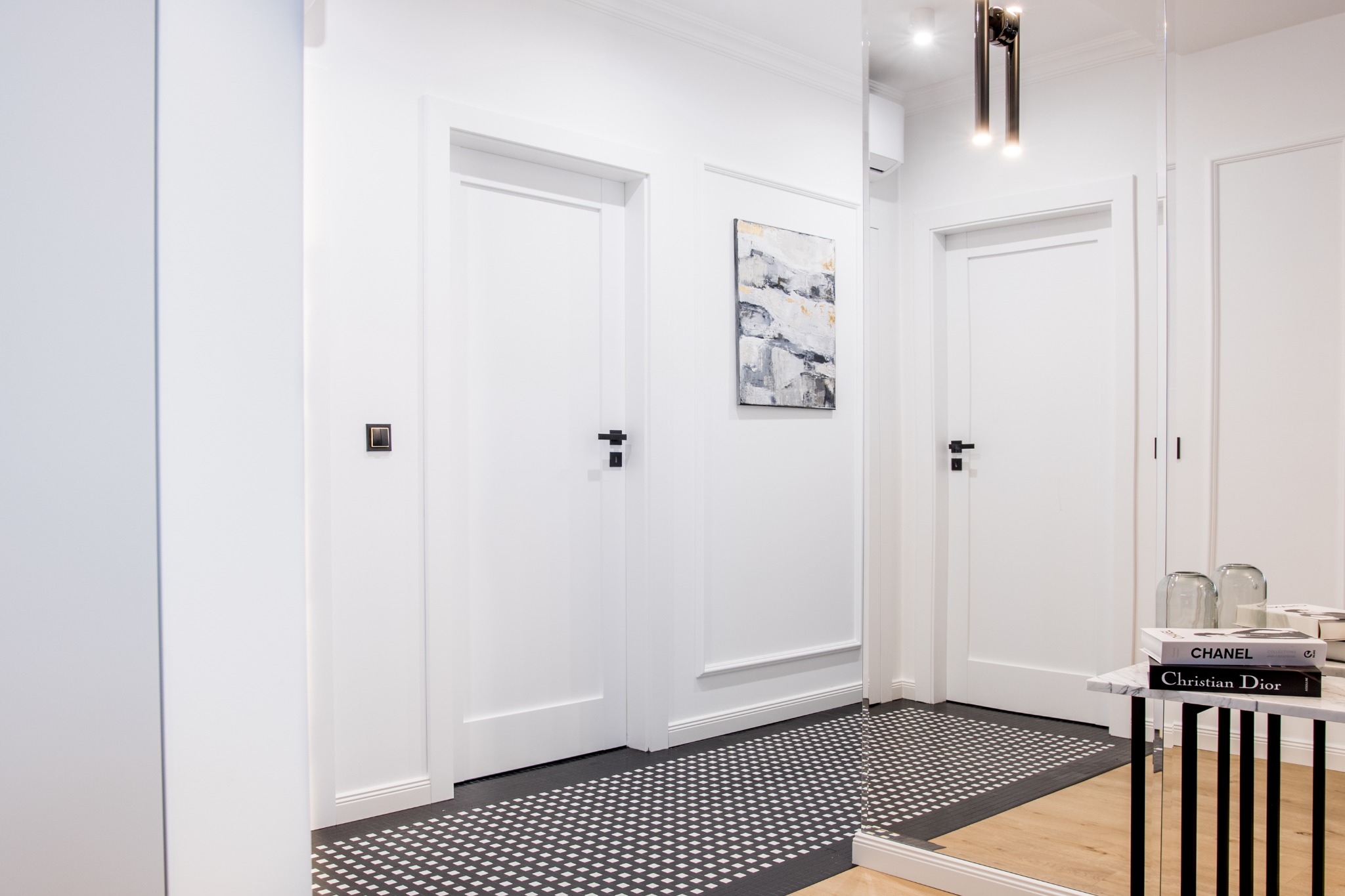 Nowoczesny przedpokój z białymi drzwiami, czarno-białą mozaiką na podłodze, abstrakcyjnym obrazem na ścianie i stolikiem z książkami Chanel i Christian Dior.