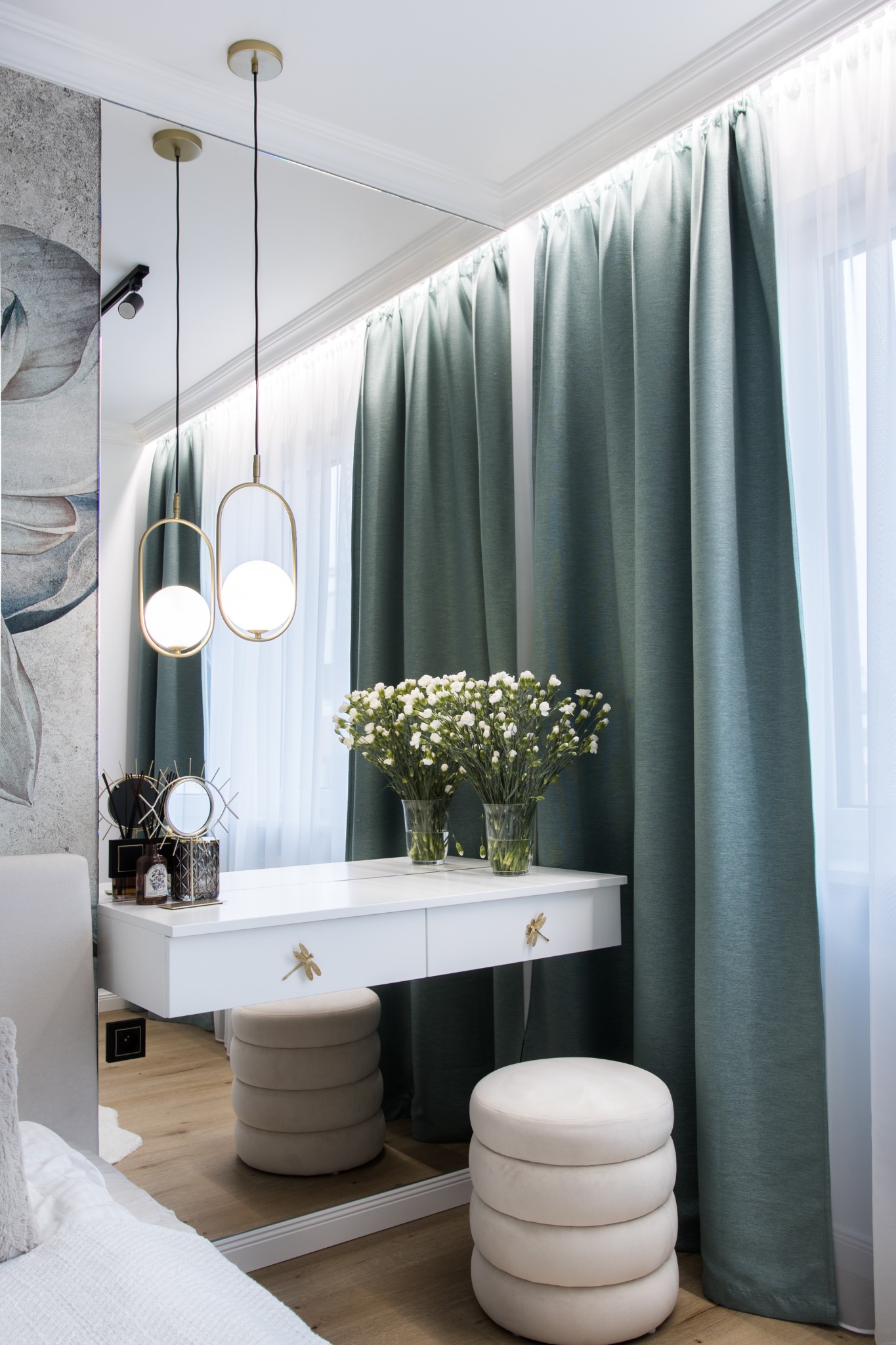 Elegancka toaletka z białym blatem, złotymi uchwytami w kształcie ważek i dwoma pufami, oświetlona wiszącymi lampami z okrągłymi kloszami, na tle zielonych zasłon i lustra.