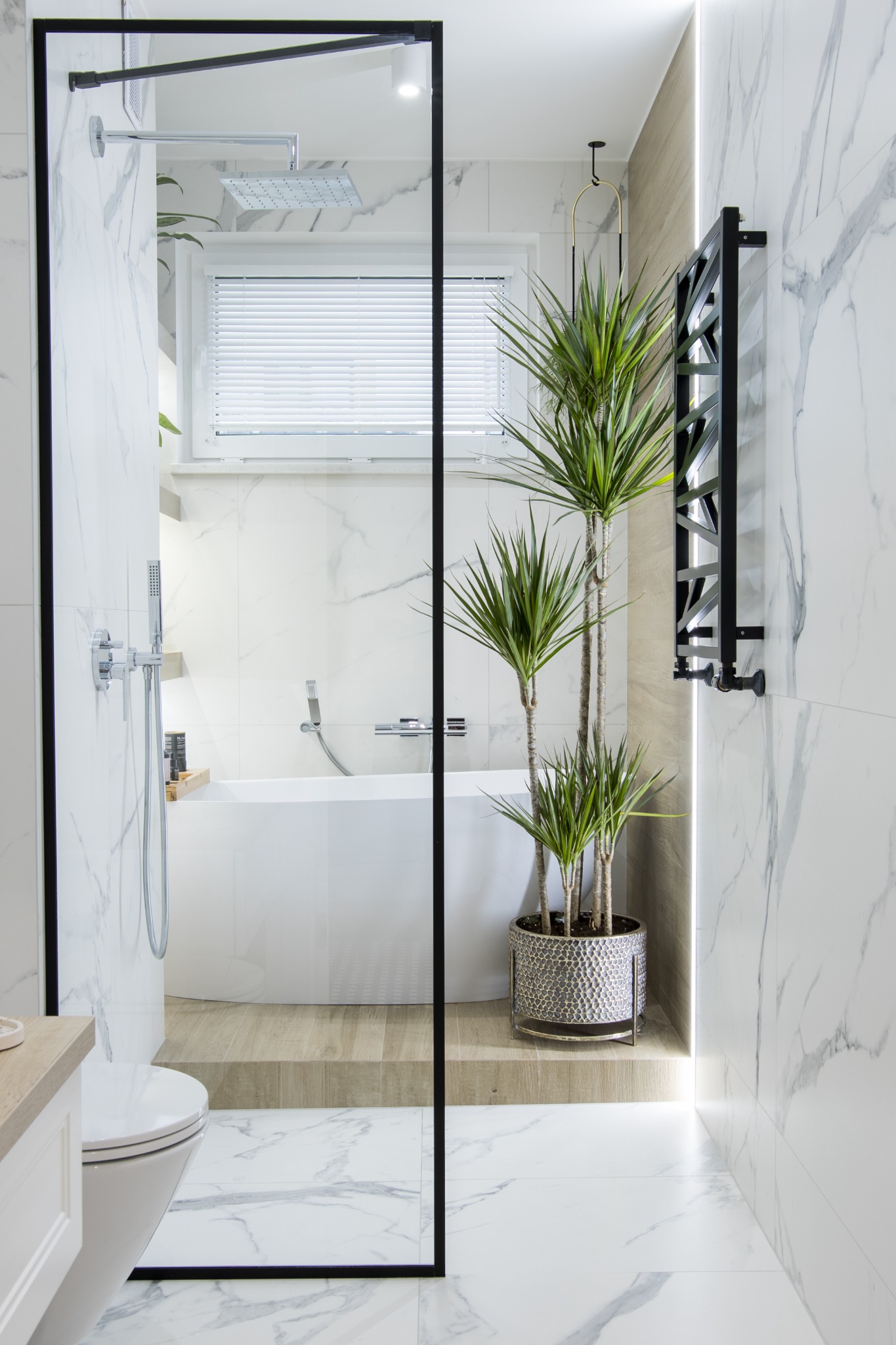 Nowoczesna łazienka z kabiną prysznicową typu walk-in, wolnostojącą wanną, grzejnikiem drabinkowym w kolorze czarnym i rośliną doniczkową w ozdobnej osłonce, wykończona białymi płytkami imitującymi...