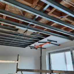 Montaż płyty gipsowo-kartonowej na metalowym ruszcie sufitowym w pomieszczeniu w Lubszy, widoczny podnośnik do płyt i fragment rusztowania.