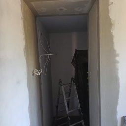 Regips - Ościeżnica po obróbce gładzią, widoczne przewody i drabina malarska w głębi. Ściana w trakcie prac wykończeniowych, szpachlowanie.