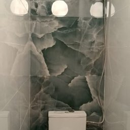 Regips - Wykończona łazienka z toaletą, ściany wyłożone płytkami imitującymi marmur w odcieniach szarości i zieleni. Nad toaletą trzy okrągłe lampy i słuchawka prysznicowa.