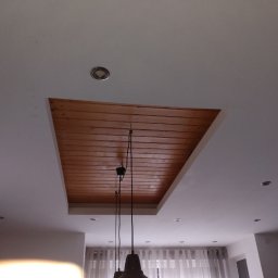 Regips - Drewniana podbitka sufitowa wpuszczona w biały sufit z oświetleniem punktowym. Widoczna część lampy wiszącej i fragment okna z firanką.