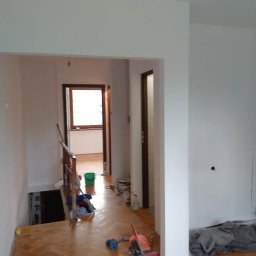 Regips - Jasny korytarz po malowaniu ścian na biało, z widocznymi narzędziami i materiałami malarskimi na parkiecie. Widok na schody i okno w tle.