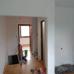 Jasny korytarz po malowaniu ścian na biało, z widocznymi narzędziami i materiałami malarskimi na parkiecie. Widok na schody i okno w tle.