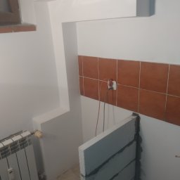 Regips - Wnętrze łazienki w trakcie remontu: murowana ścianka z bloczków, brązowe płytki na ścianie, grzejnik. Widoczne przewody elektryczne. Prace murarskie i instalacyjne.