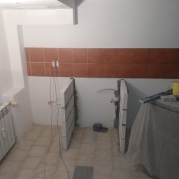 Regips - Wnętrze łazienki w trakcie remontu: widoczne ściany z brązowymi płytkami, białe ściany, fragment grzejnika i  konstrukcja z bloczków betonowych pod umywalkę.