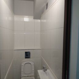 Regips - Biała, wąska toaleta po remoncie z umywalką i WC. Ściany wyłożone płytkami z delikatnym pionowym wzorem. Widoczny fragment ciemnej framugi drzwi.