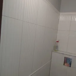 Wykończona ściana w łazience z białymi, pionowo żłobionymi płytkami i dekoracyjną listwą. Widoczny fragment toalety i butelka środka czyszczącego.