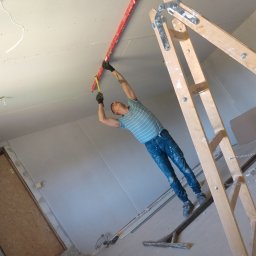 Regips - Pracownik na drabinie mierzy poziom sufitu z płyty karton-gips, używając poziomicy i miarki. Widoczne niedokończone łączenia płyt i instalacja elektryczna.