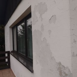 Regips - Narożnik budynku z łuszczącą się, odpadającą farbą na elewacji, widoczne okno z ciemną ramą i fragment balkonu z brązową barierką. Zły stan powłoki malarskiej.