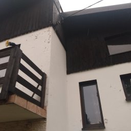 Regips - Narożne ujęcie budynku z balkonem i łuszczącą się farbą na elewacji, widoczne okna z ciemnymi ramami, oświetlenie zewnętrzne i fragment dachu.