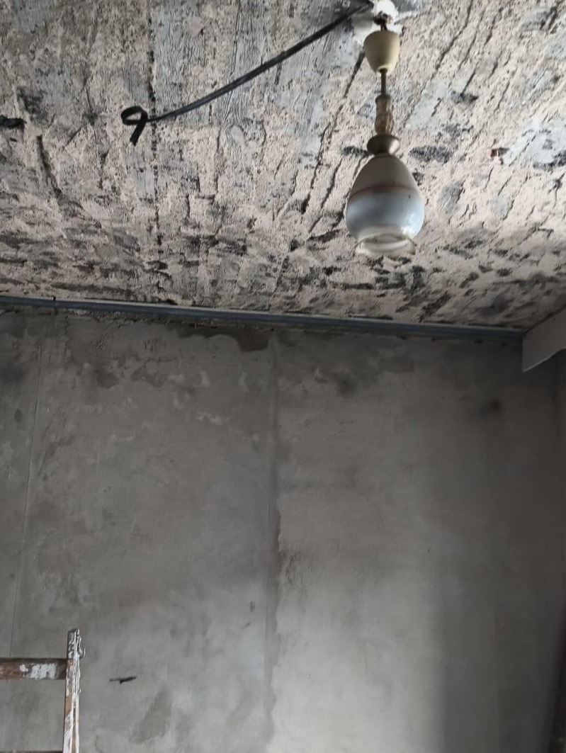 Surowa ściana i nierówny sufit z lampą wiszącą. Widoczne ślady po instalacji i przygotowaniu do dalszych prac wykończeniowych. Fragment drabiny malarskiej w rogu.