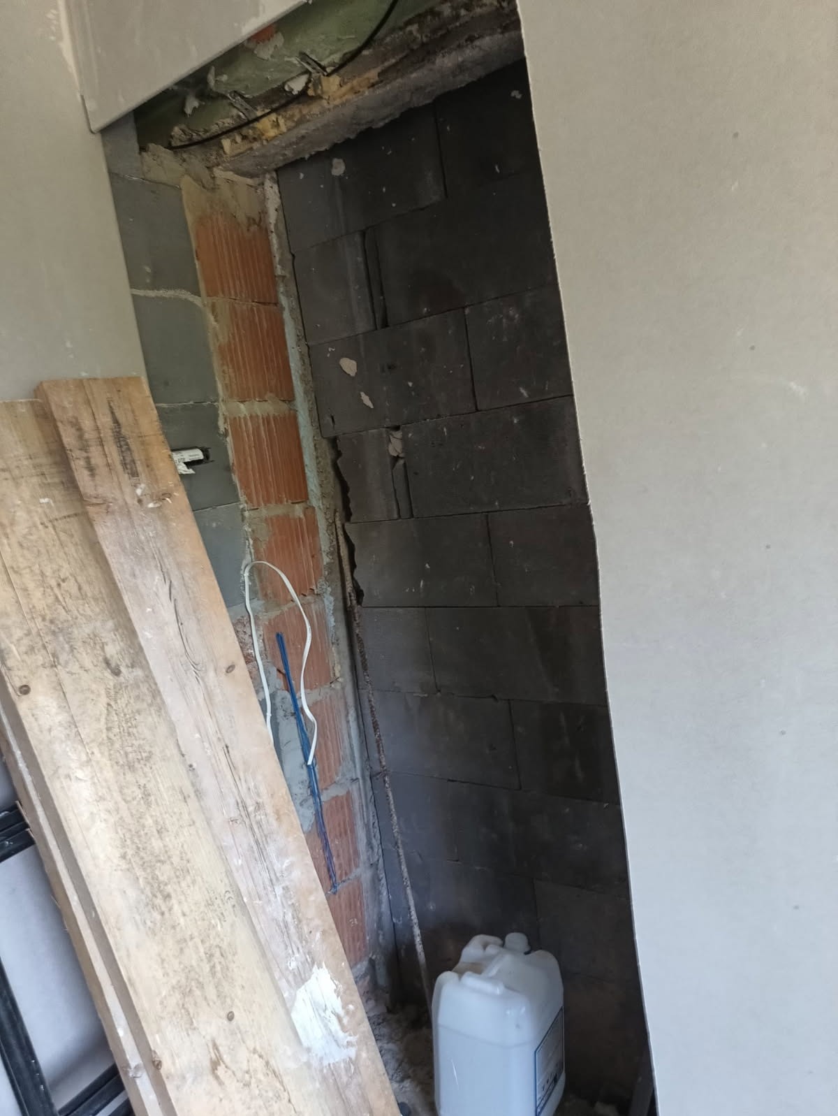 Wnęka w ścianie z widoczną konstrukcją z cegieł i bloczków, częściowo zakryta płytą gipsową. Obok leżą deski i pojemnik z płynem, sugerujące prace remontowe.