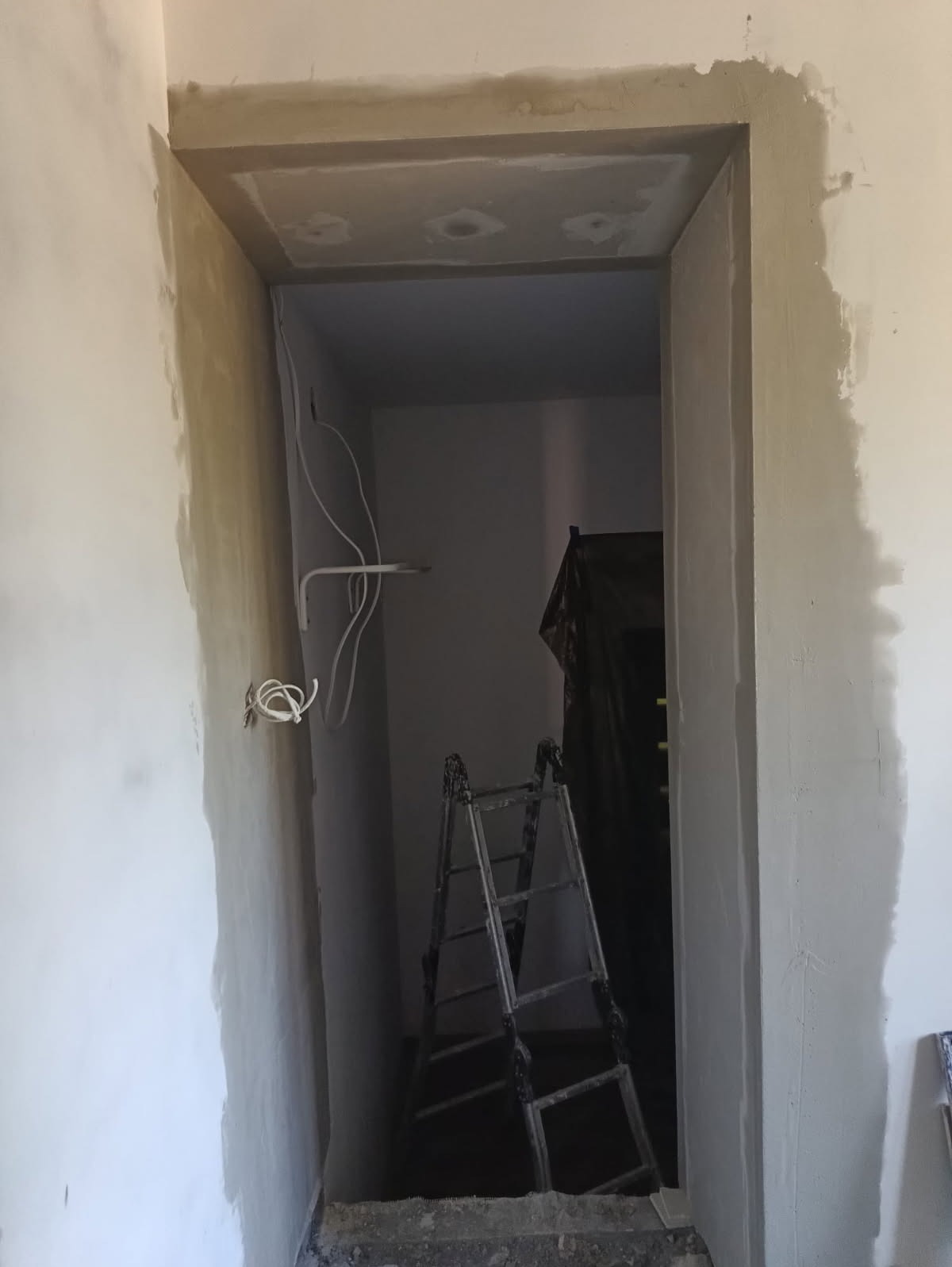 Ościeżnica po obróbce gładzią, widoczne przewody i drabina malarska w głębi. Ściana w trakcie prac wykończeniowych, szpachlowanie.