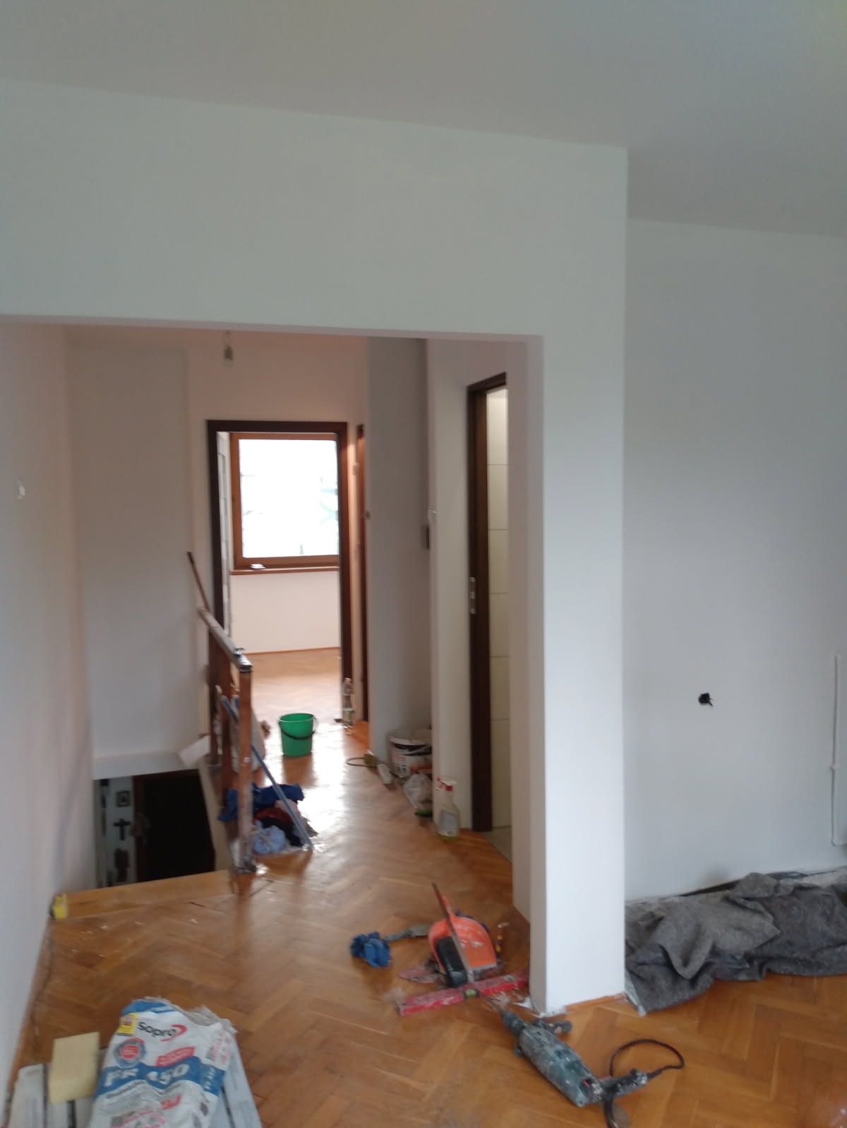 Wnętrze po malowaniu: białe ściany, drewniana podłoga, narzędzia malarskie i resztki materiałów budowlanych na podłodze. Widok na korytarz i otwarte drzwi.