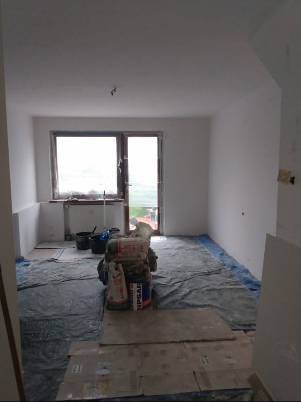 Pokój w trakcie remontu: zabezpieczona podłoga, worki z gipsem, wiadra. Widoczne przygotowania do prac wykończeniowych, ściany przygotowane do malowania.