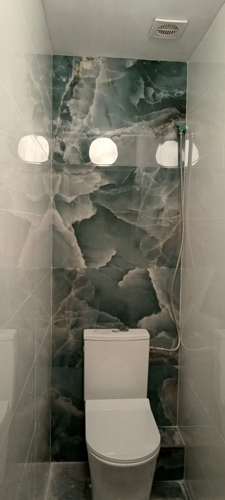 Wykończona łazienka z toaletą, ściany wyłożone płytkami imitującymi marmur w odcieniach szarości i zieleni. Nad toaletą trzy okrągłe lampy i słuchawka prysznicowa.