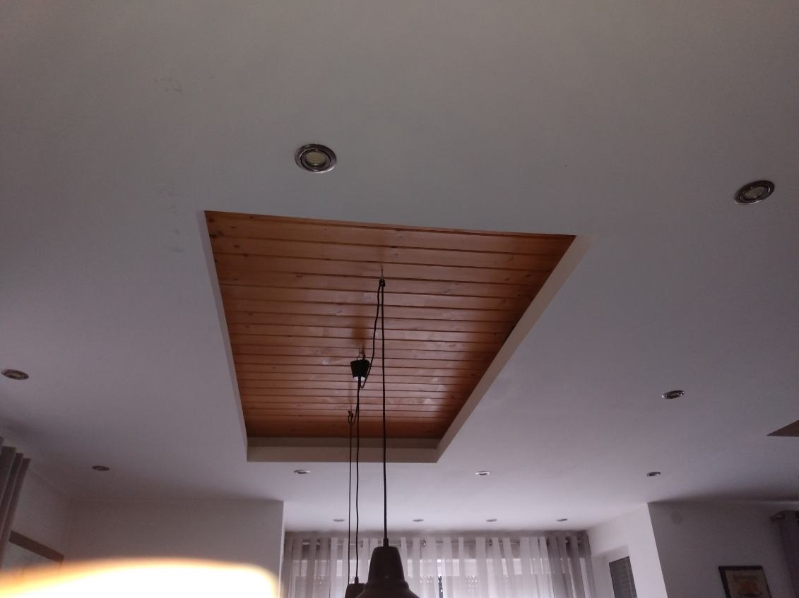 Drewniana podbitka sufitowa wpuszczona w biały sufit z oświetleniem punktowym. Widoczna część lampy wiszącej i fragment okna z firanką.