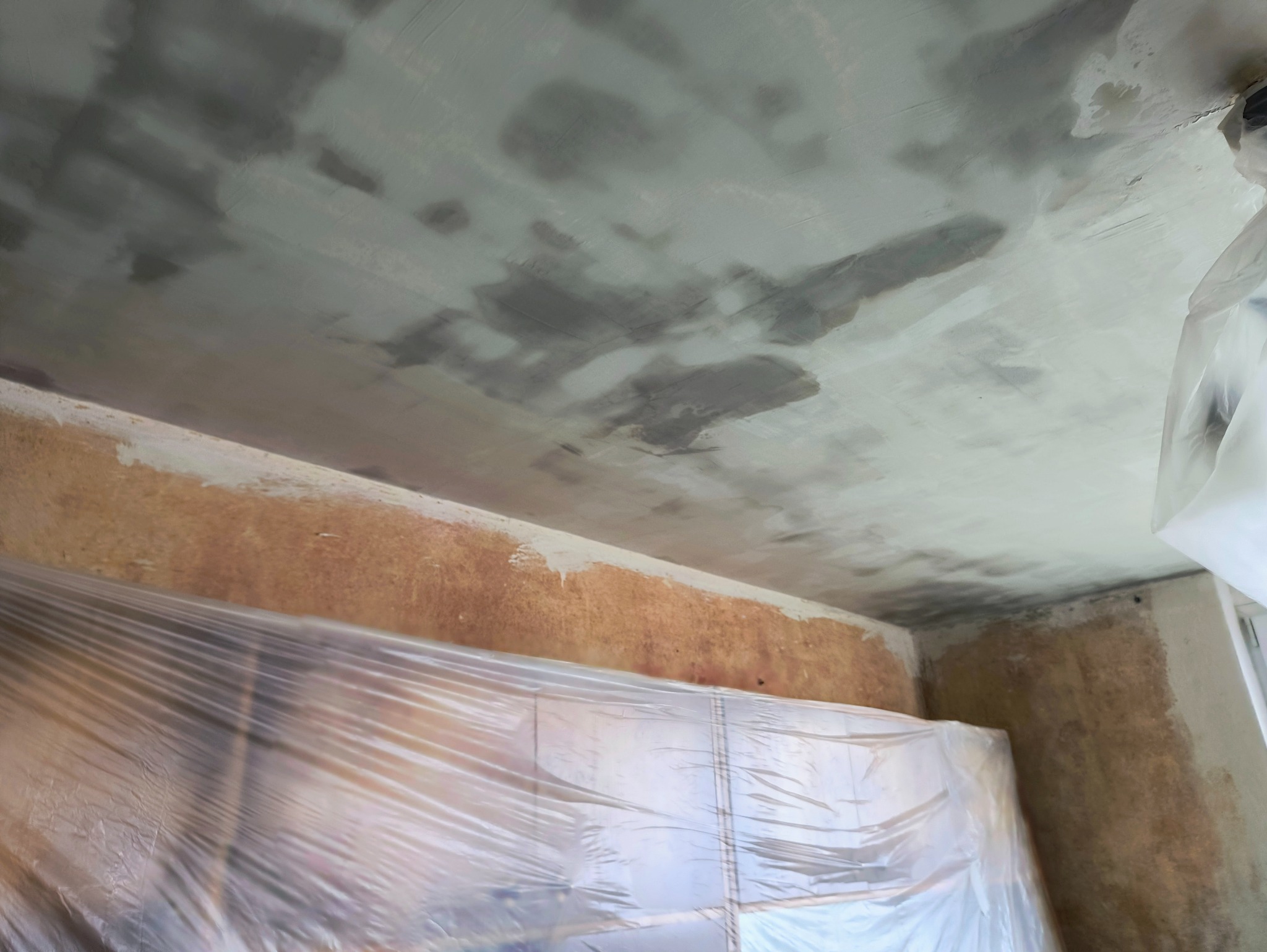 Niedokończony sufit po nałożeniu gładzi, widoczne mokre plamy. Okno zabezpieczone folią malarską, przygotowanie do dalszych prac.