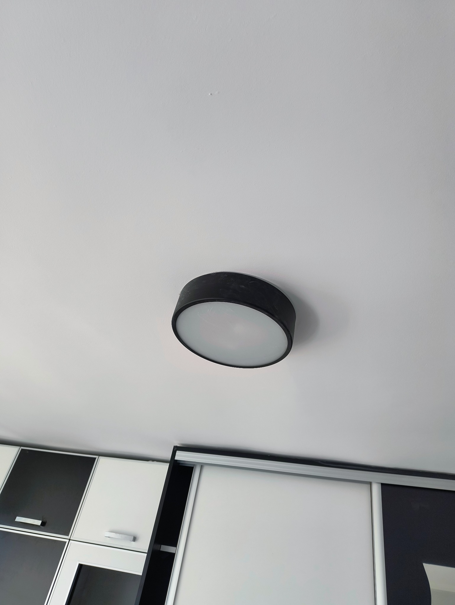 Zamontowana okrągła lampa sufitowa w nowoczesnym wnętrzu. Widoczne białe ściany i fragment szafy przesuwnej z czarno-białymi frontami.
