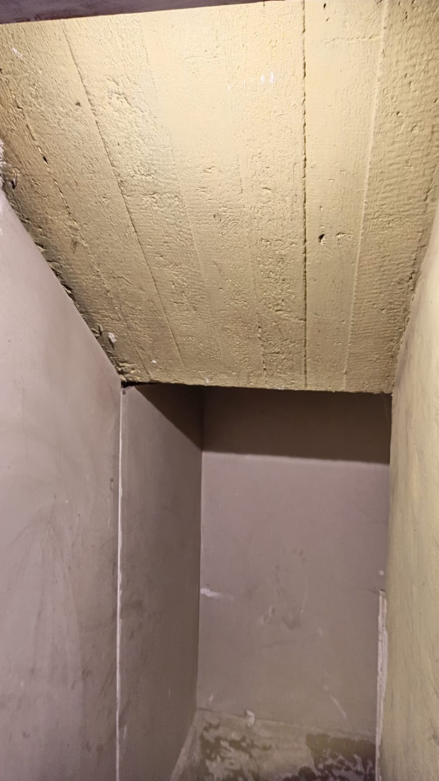 Narożnik pomieszczenia z surowym, beżowym sufitem z desek i świeżo otynkowanymi ścianami w odcieniach szarości i beżu. Widoczne niedoskonałości i nierówności powierzchni.