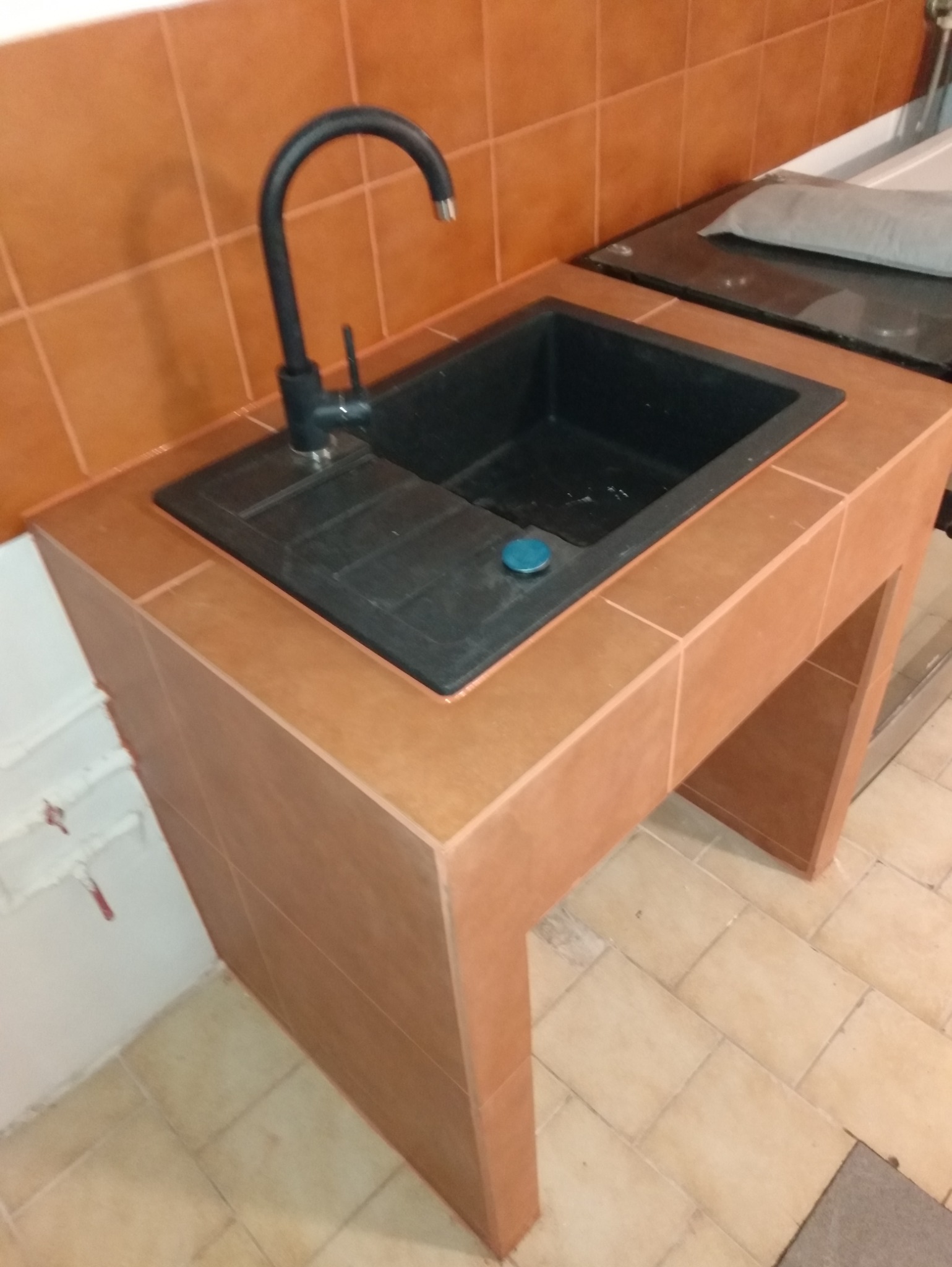 Wykończony zlew z czarną armaturą i brązowymi płytkami, zabudowany w sposób minimalistyczny. Widoczne rury i podłączenia. Ukończona realizacja w łazience.