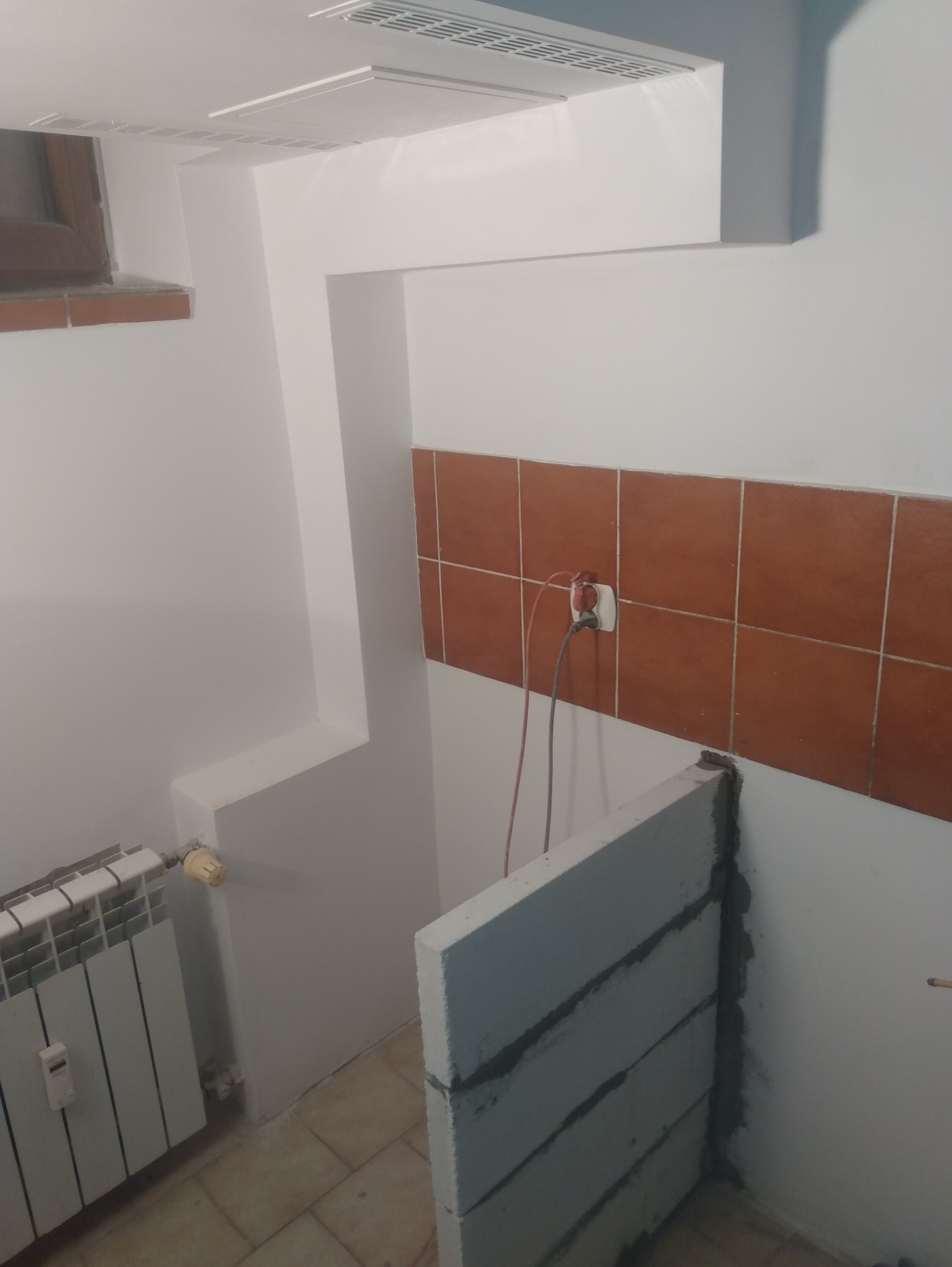 Wnętrze łazienki w trakcie remontu: murowana ścianka z bloczków, brązowe płytki na ścianie, grzejnik. Widoczne przewody elektryczne. Prace murarskie i instalacyjne.