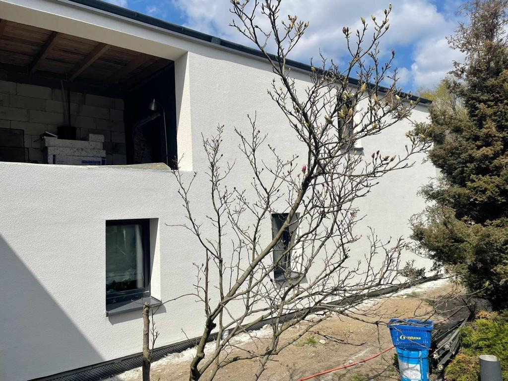 Biała elewacja domu w trakcie budowy, widoczne okno, fragment niezadaszonego piętra z materiałami budowlanymi i drzewo magnolii na pierwszym planie