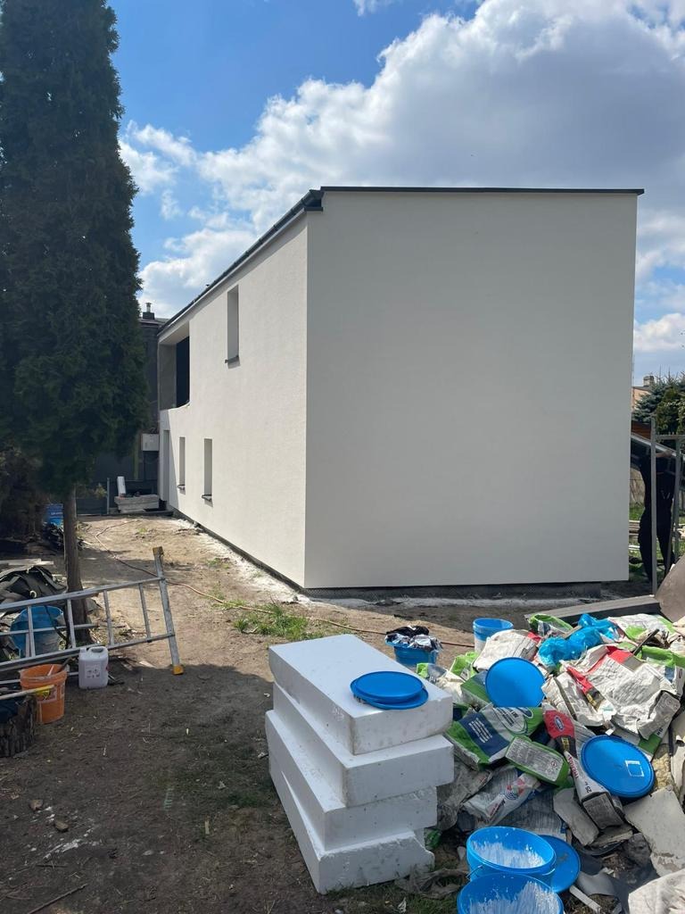 Dwukondygnacyjny dom z jasną elewacją, widok z boku na niezagospodarowany teren z materiałami budowlanymi i drabiną.