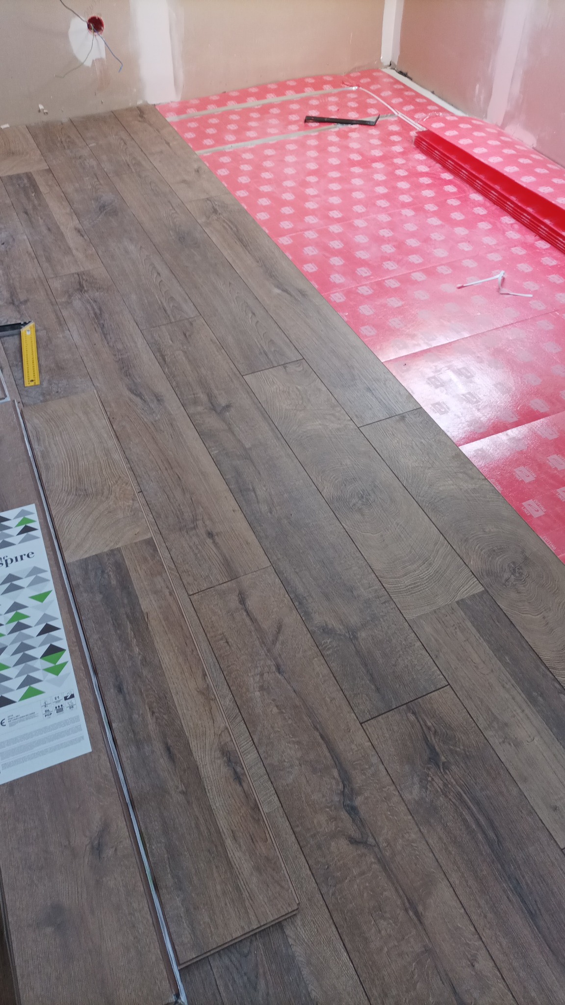 Układanie paneli podłogowych w pomieszczeniu, widoczna izolacja pod panele w kolorze różowym, poziomica i opakowanie paneli z logo 'Spire'.