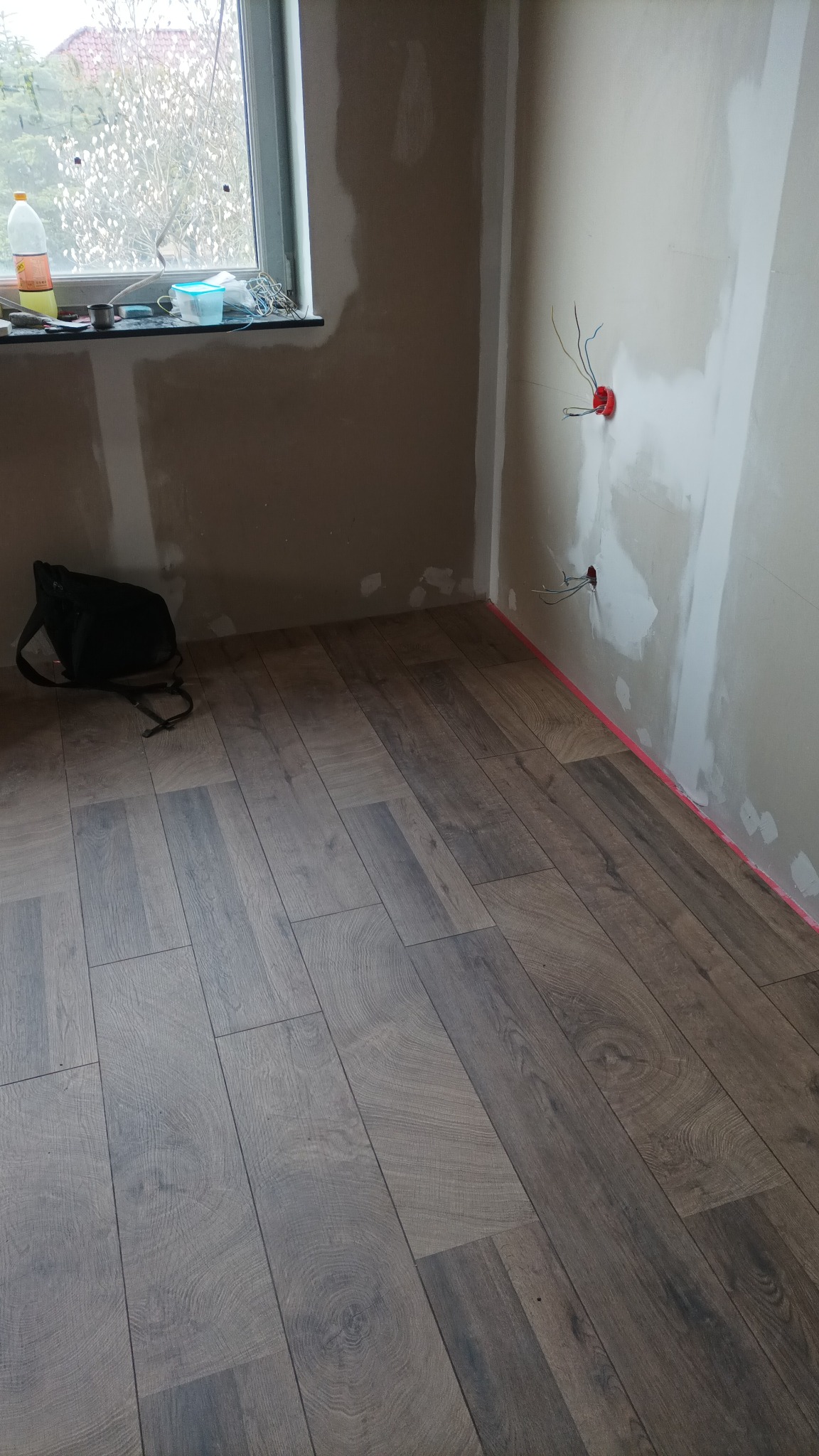 Układanie paneli podłogowych w nowym wnętrzu, widoczne surowe ściany z instalacją elektryczną i torba narzędziowa przy oknie.