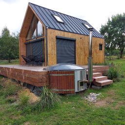 Studio Tarasów Mariusz Szupiluk - Nowoczesny domek z drewnianą elewacją, roletami zewnętrznymi i metalowym dachem, otoczony tarasem z bali oraz zewnętrzną balią z piecem, w tle zielony trawnik i drzewa.