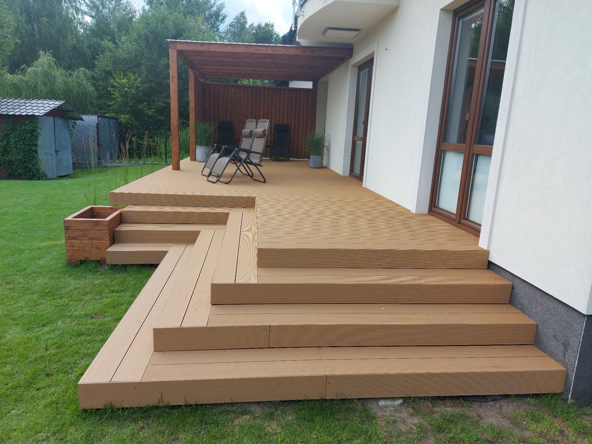 Nowoczesny taras z desek kompozytowych w odcieniach brązu, z niskimi schodami prowadzącymi do trawnika, zadaszony drewnianą pergolą, na której stoją dwa krzesła ogrodowe, z widokiem na zielony ogród.