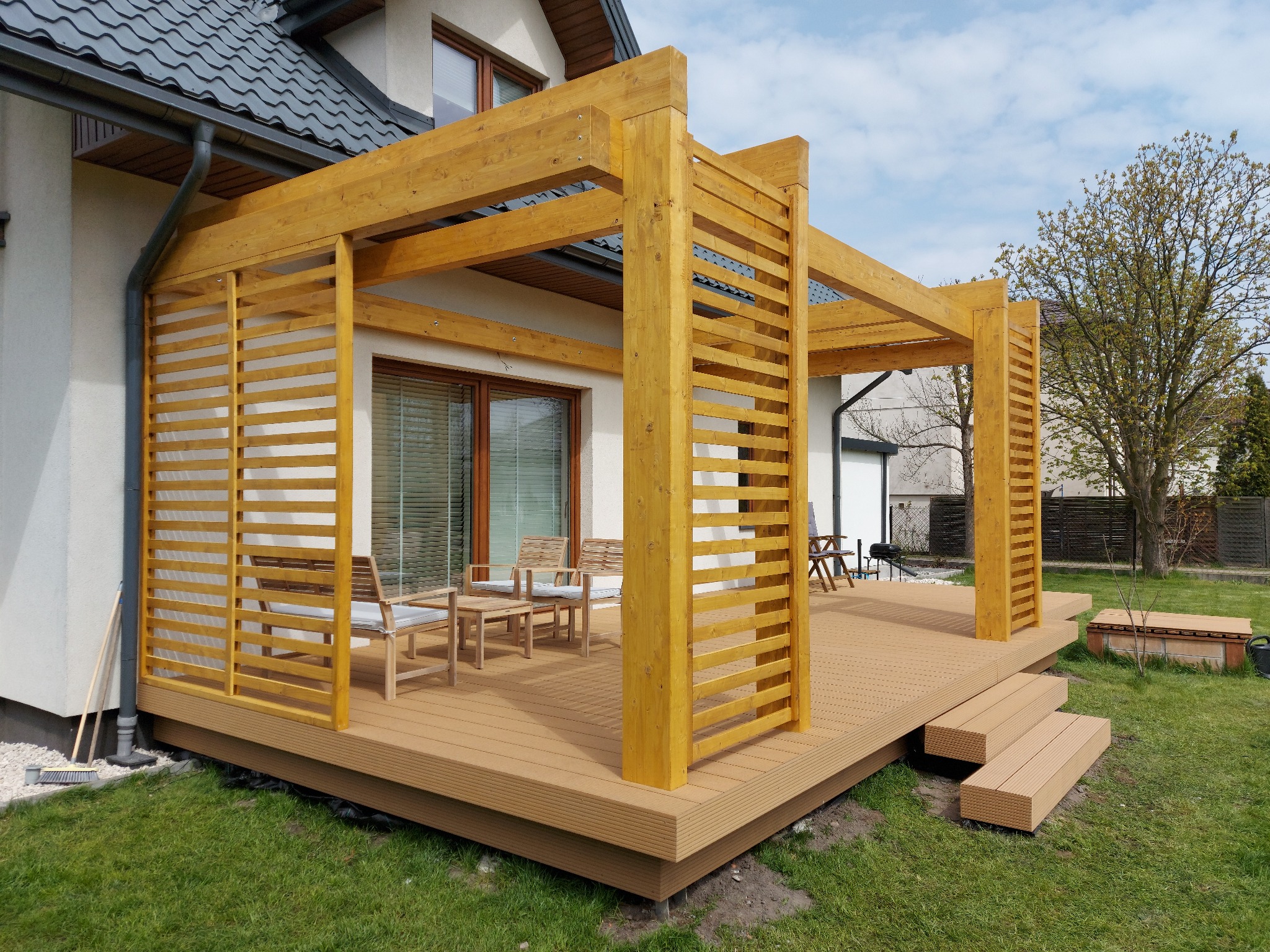 Jasna pergola z drewnianymi lamelami, połączona z tarasem z desek kompozytowych, zestaw mebli ogrodowych z jasnymi poduszkami, widok na trawnik i dom.