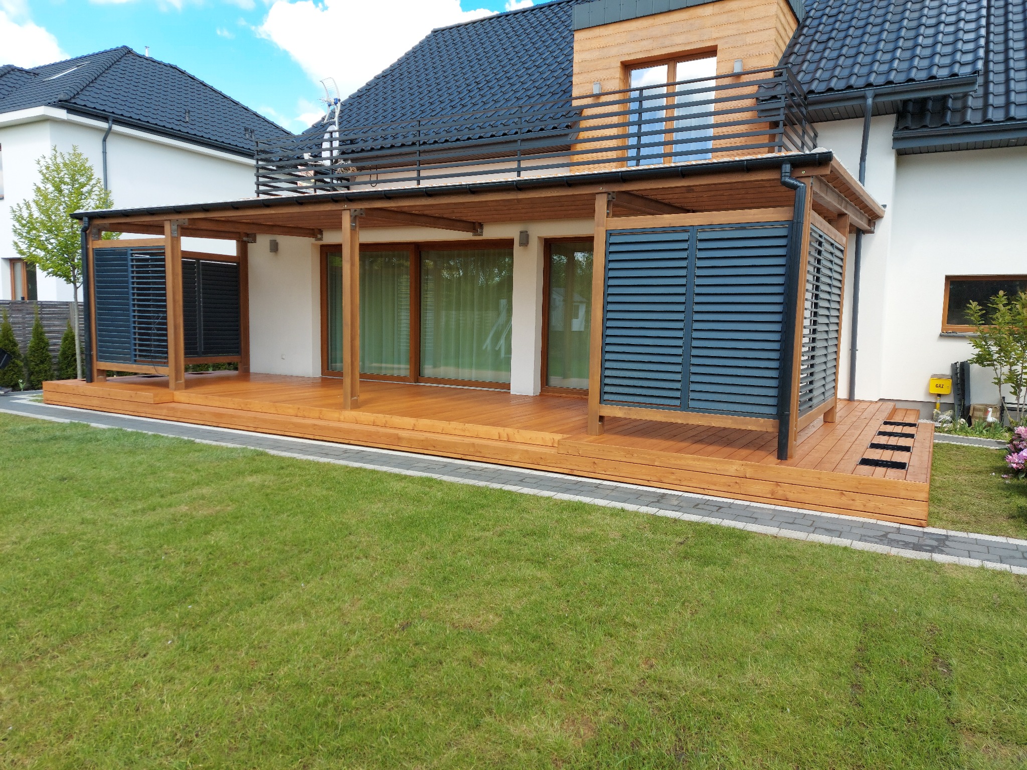 Zadaszony taras z drewnianą podłogą i panelami żaluzjowymi, przylegający do nowoczesnego domu z drewnianą elewacją na piętrze, otoczony zielonym trawnikiem.