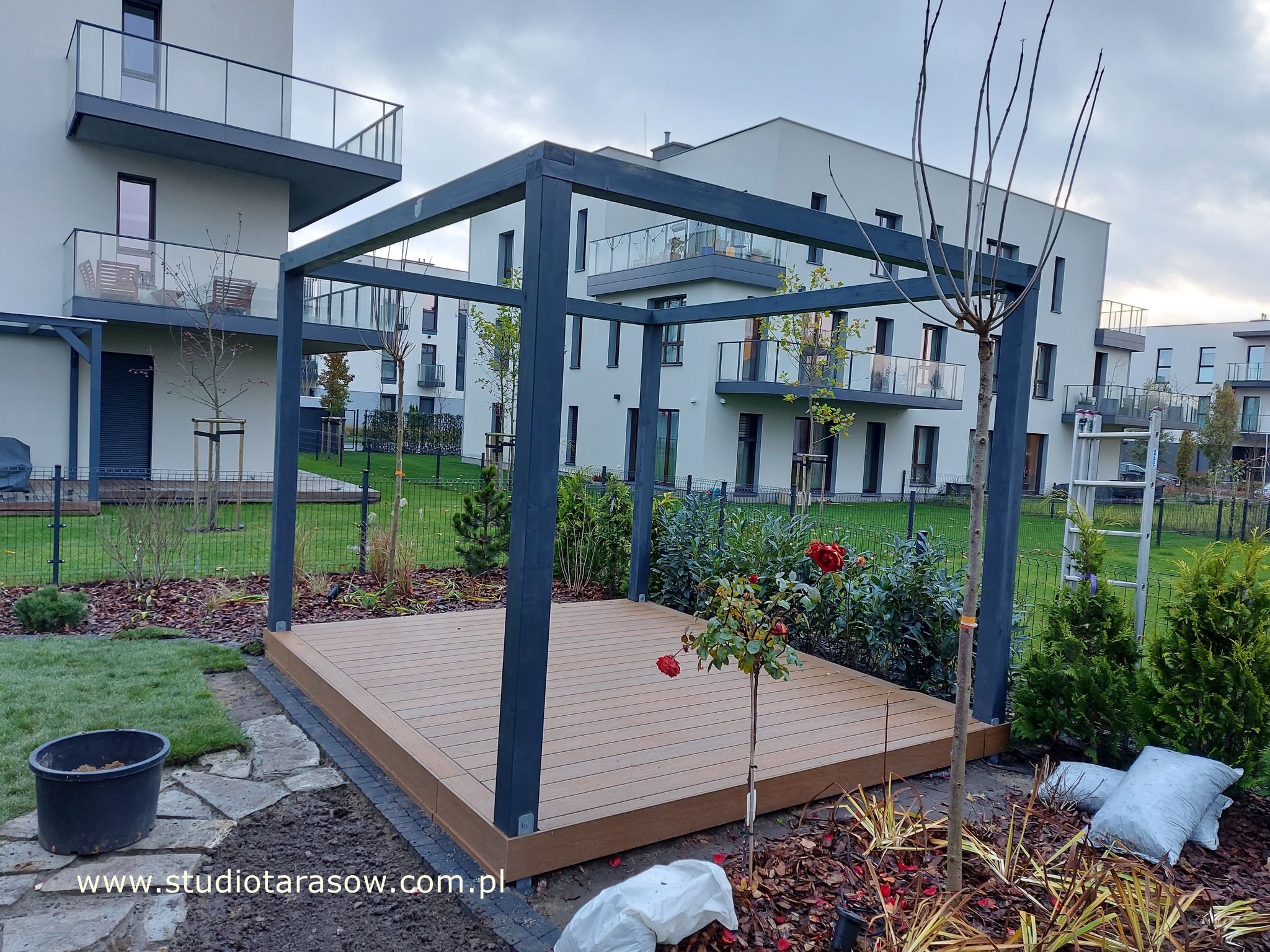 Nowoczesna pergola z ciemną ramą na tarasie z desek w kolorze jasnego drewna, otoczona zielenią i elementami krajobrazu, z budynkami mieszkalnymi w tle.