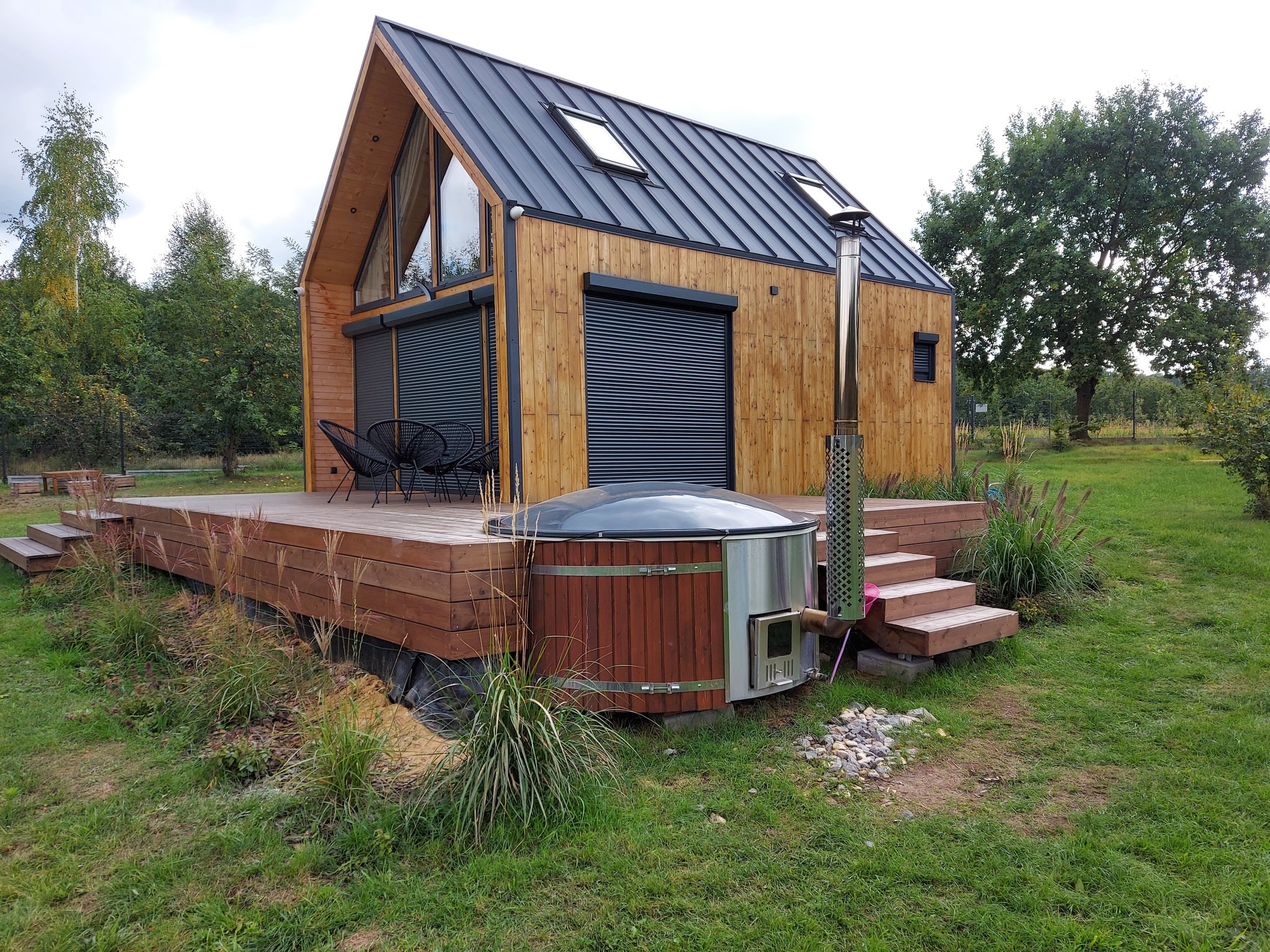 Nowoczesny domek z drewnianą elewacją, roletami zewnętrznymi i metalowym dachem, otoczony tarasem z bali oraz zewnętrzną balią z piecem, w tle zielony trawnik i drzewa.