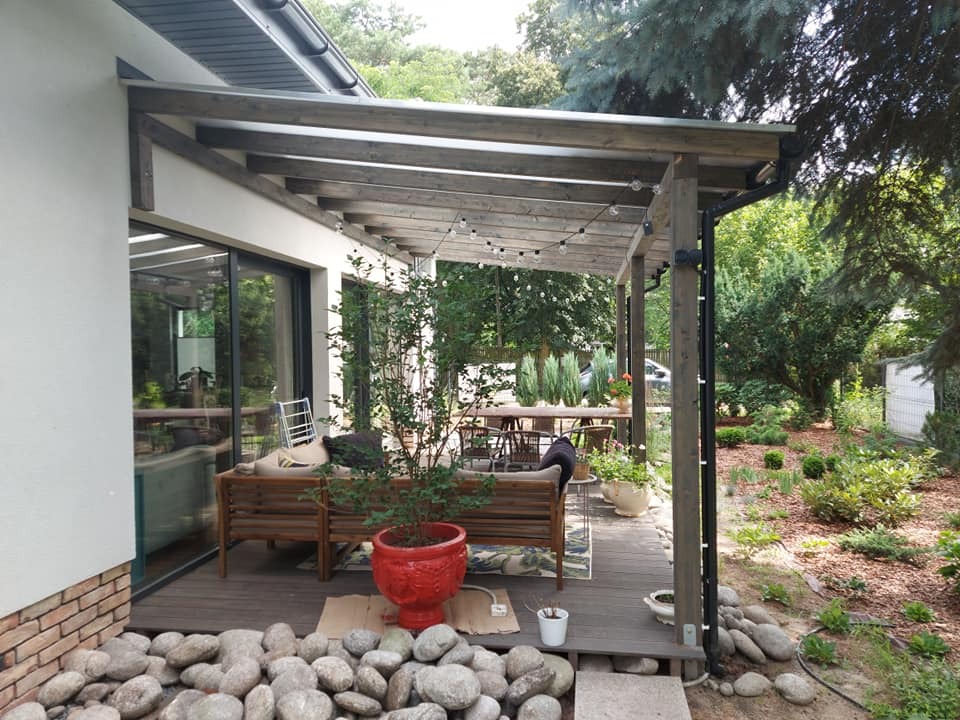 Zadaszony taras z drewnianą konstrukcją, oświetlony girlandami, z widokiem na zadbany ogród, w tle stół i krzesła.