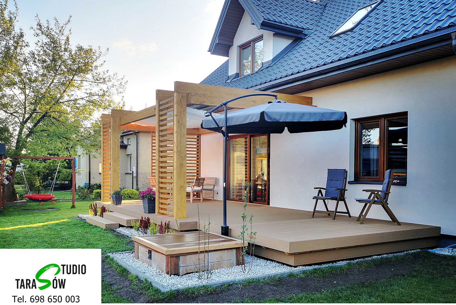 Nowoczesny taras z desek w kolorze jasnego drewna, z drewnianą pergolą, parasolem i meblami ogrodowymi, otoczony zielonym trawnikiem i roślinnością, widok na dom z ciemną dachówką.