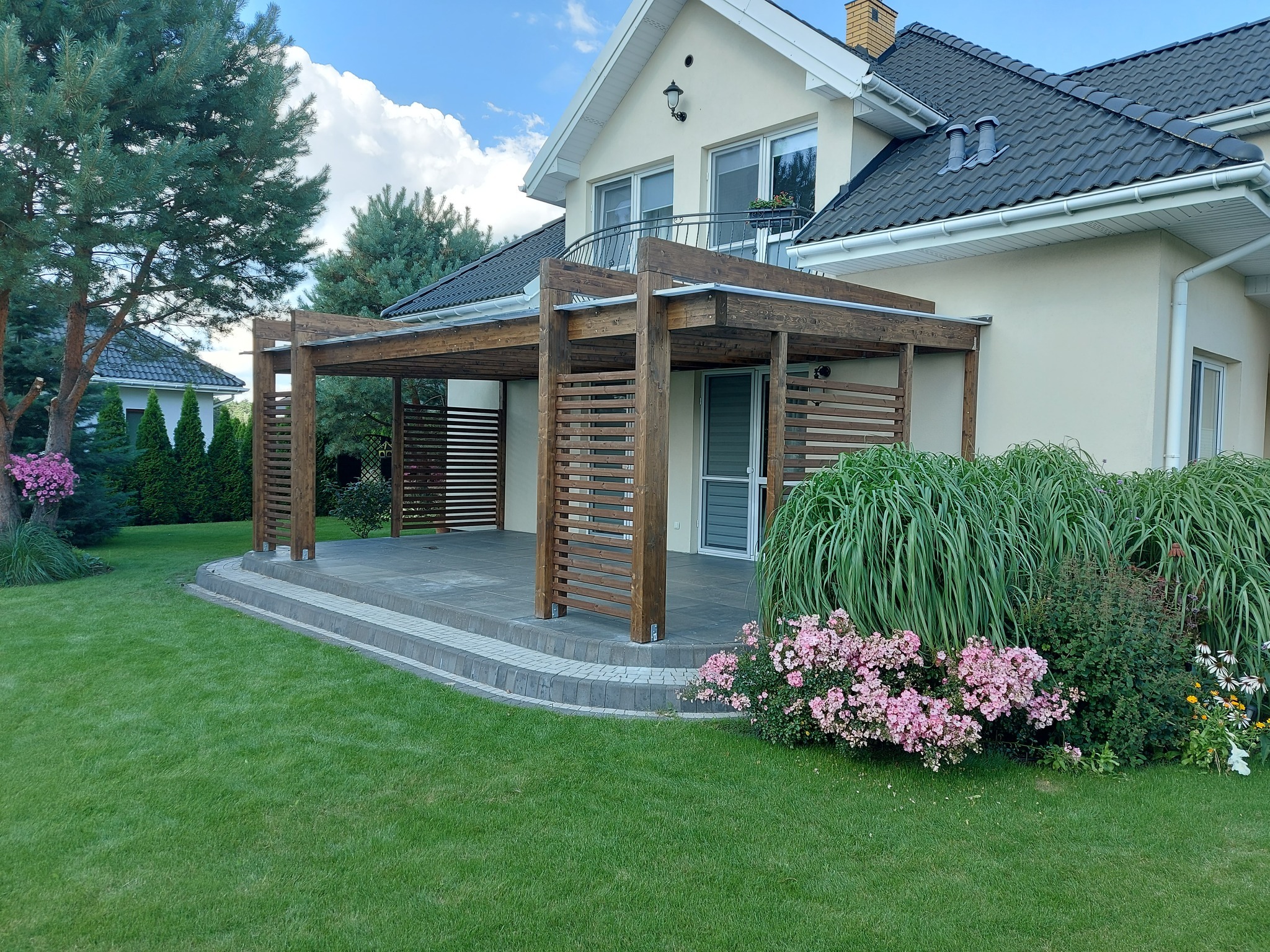 Drewniana pergola z poziomymi listwami, przylegająca do domu z balkonem, otoczona zielonym trawnikiem i kwitnącymi krzewami.