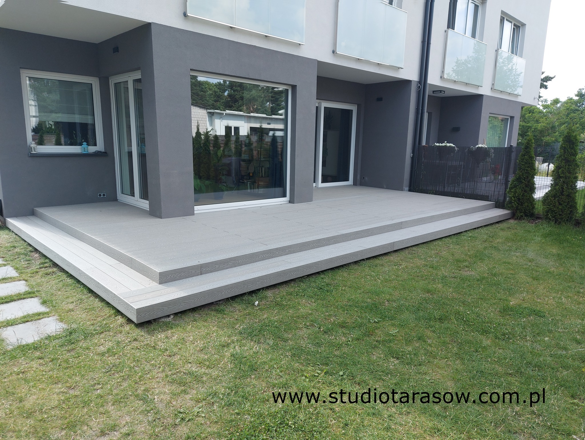 Szara, dwupoziomowa konstrukcja tarasowa z kompozytu, przylegająca do nowoczesnego domu z dużymi oknami, widziana z poziomu trawnika.