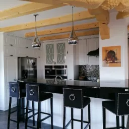 Nowoczesna kuchnia z białymi szafkami, czarnym blatem i hokerami z czarnej tkaniny z ozdobnymi srebrnymi nitami, drewniane belki stropowe z rzeźbieniami winogron.