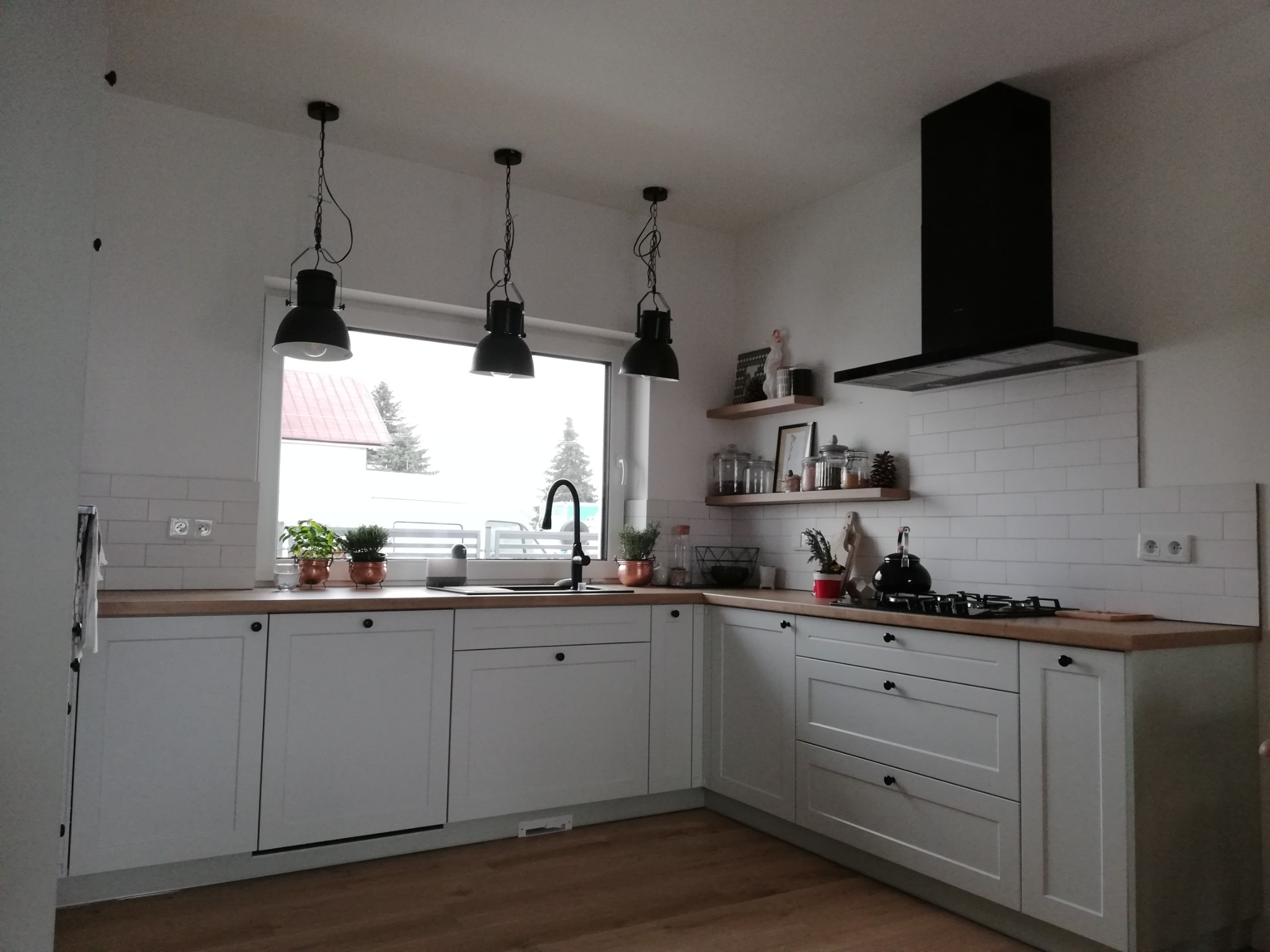 Jasna kuchnia z drewnianym blatem, białymi szafkami z ciemnymi uchwytami, czarną armaturą i trzema wiszącymi lampami w stylu industrialnym. Na półkach dekoracje i akcesoria kuchenne.