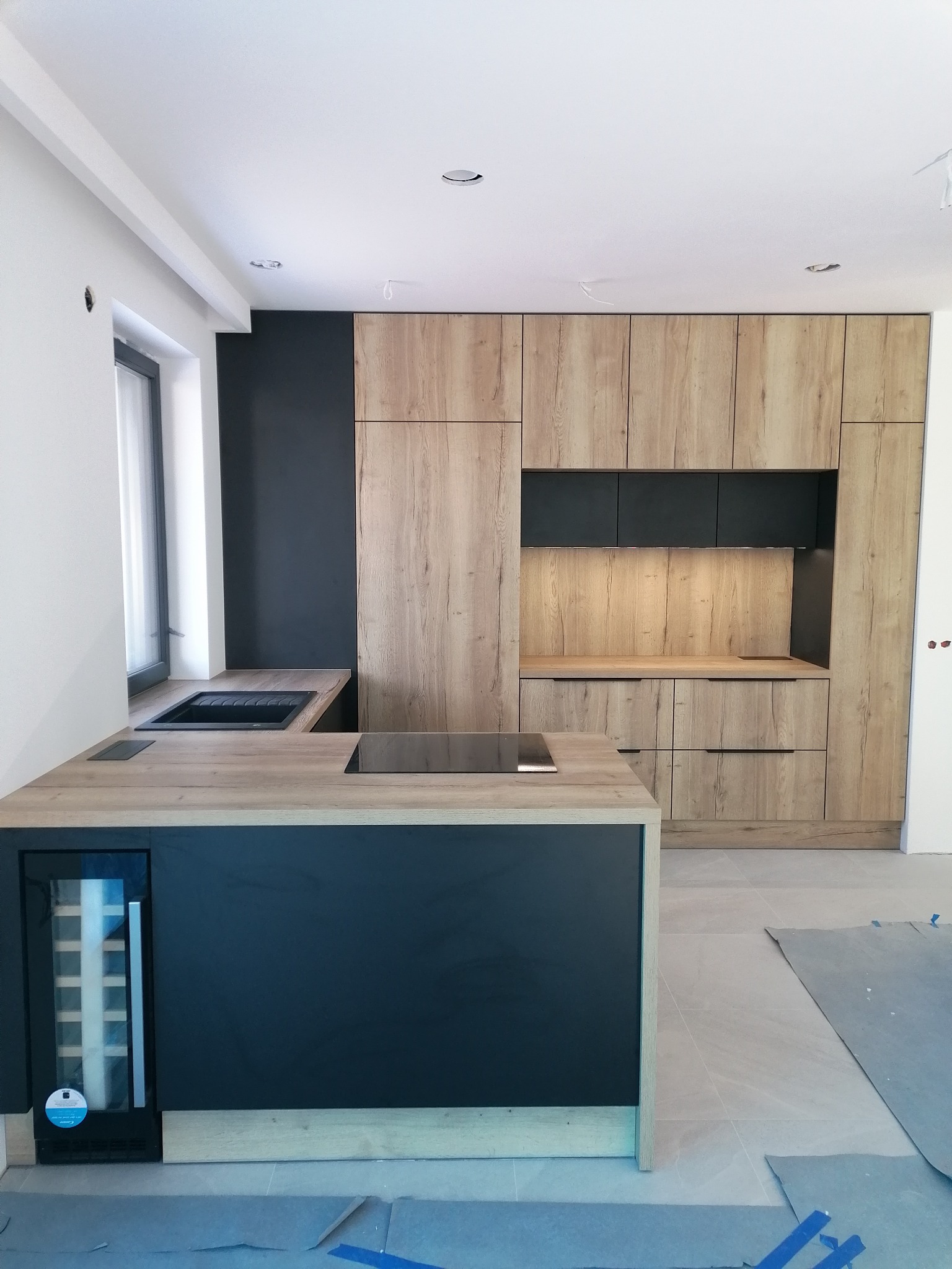 Nowoczesna kuchnia z drewnianymi frontami szafek i czarnym blatem wyspy kuchennej z wbudowaną chłodziarką na wino, widoczna część pomieszczenia w trakcie wykańczania.